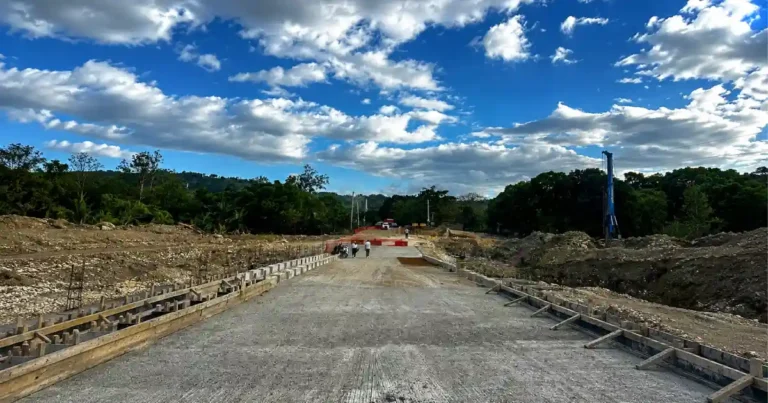 Toma panorámica que muestra el desvío provisional en el río Camú, en Yásica. En primer plano, hay un gran letrero naranja de construcción con la palabra 'DESVIO' en letras negras y una excavadora amarilla activa trabajando en el terreno rocoso del río.
